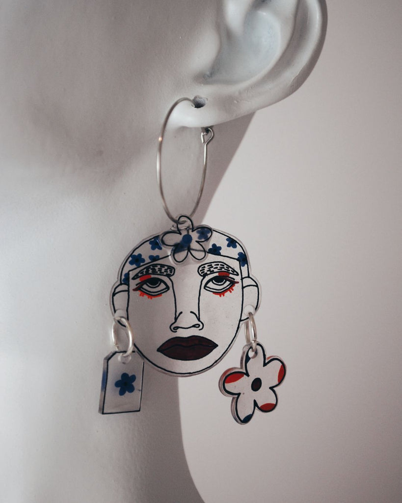 Foto van  één van de paar oorbellen gepresenteerd aan een witte buste , tegen een witte achtergrond. De oorbellen zijn 2 doorschijnende gezichtjes met bloemen details in paars en oranje 