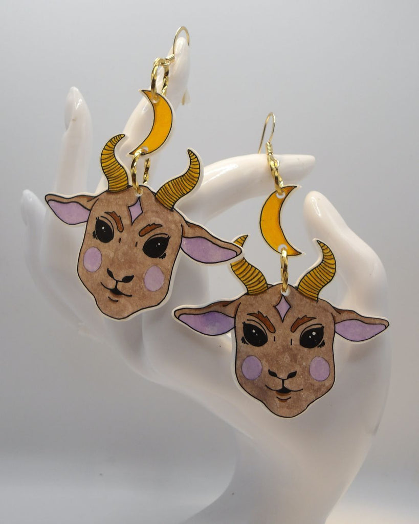 Foto van een paar oorbellen in halloween thema.  De oorbellen hebben goudkleurige hardware, en een tekening van een schattig duivels geitje, hangend aan een maan. De kleuren zijn beige, roze en geel. 