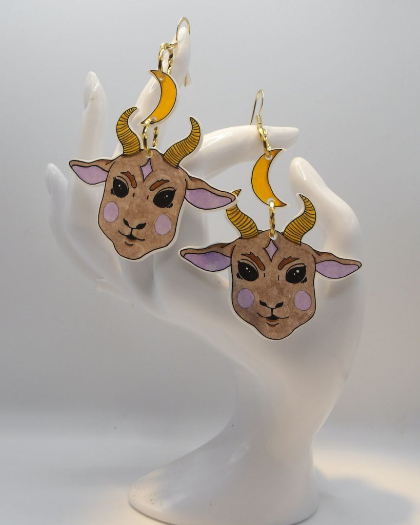 Foto van een paar oorbellen in halloween thema.  De oorbellen hebben goudkleurige hardware, en een tekening van een schattig duivels geitje, hangend aan een maan. De kleuren zijn beige, roze en geel. 