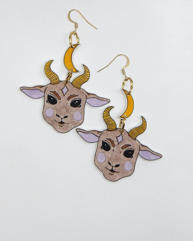 Foto van een paar oorbellen in halloween thema.  De oorbellen hebben goudkleurige hardware, en een tekening van een schattig duivels geitje, hangend aan een maan. De kleuren zijn beige, roze en geel. 