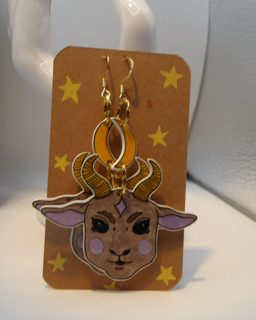 Foto van een paar oorbellen in halloween thema.  De oorbellen hebben goudkleurige hardware, en een tekening van een schattig duivels geitje, hangend aan een maan. De kleuren zijn beige, roze en geel. 