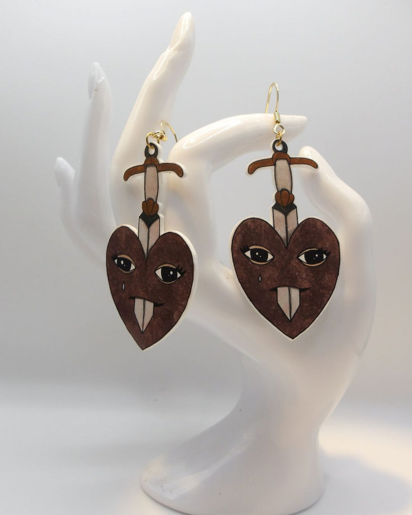 Foto van een paar oorbellen in halloween thema.  De oorbellen hebben goudkleurige hardware, en een tekening van hart dat doorboord wordt door een zwaard