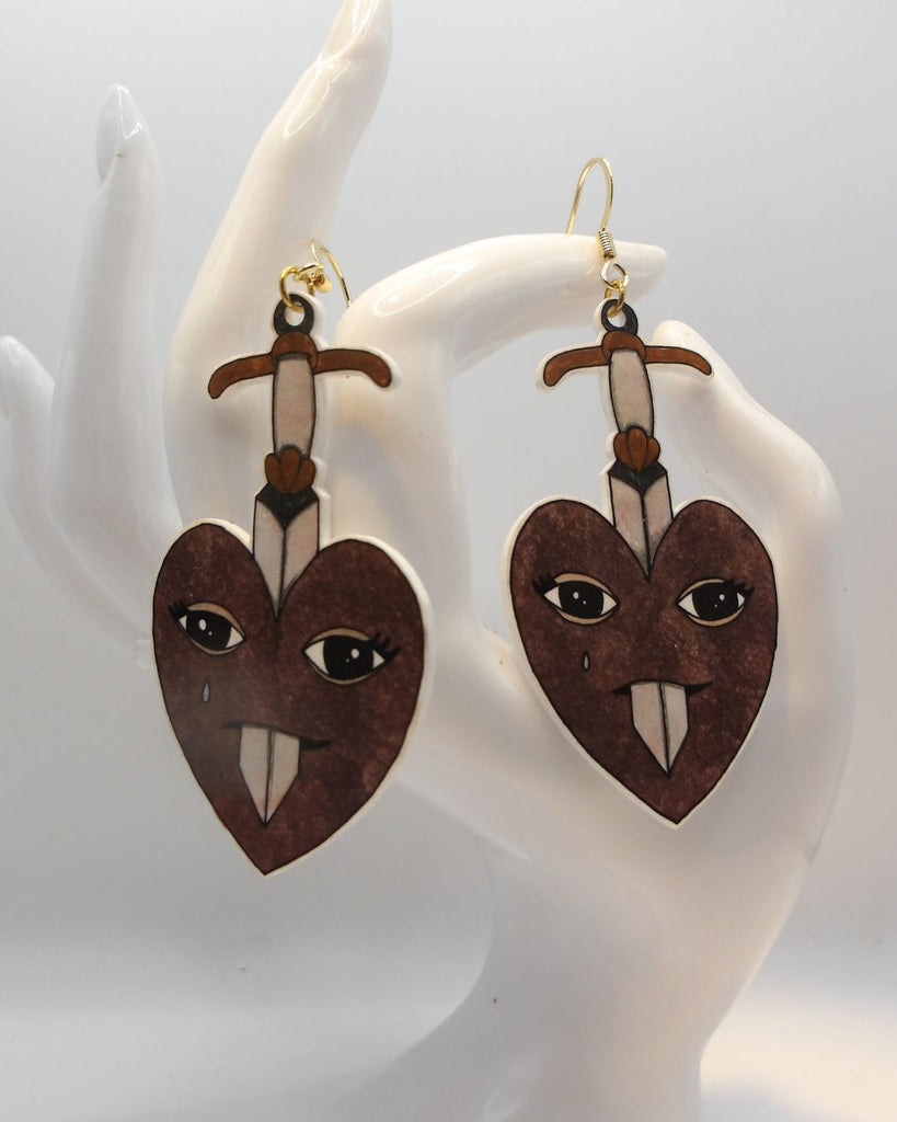 Foto van een paar oorbellen in halloween thema.  De oorbellen hebben goudkleurige hardware, en een tekening van hart dat doorboord wordt door een zwaard