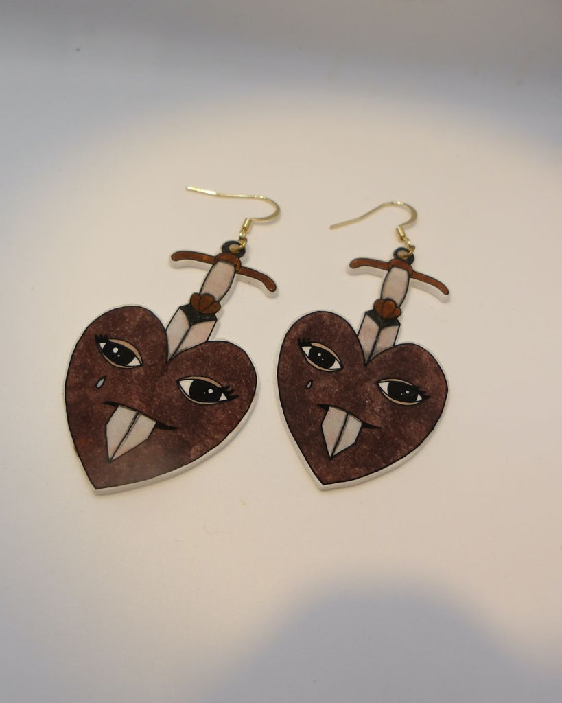 Foto van een paar oorbellen in halloween thema.  De oorbellen hebben goudkleurige hardware, en een tekening van hart dat doorboord wordt door een zwaard