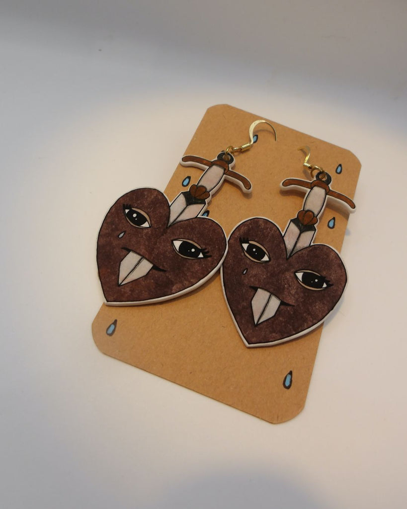 Foto van een paar oorbellen in halloween thema.  De oorbellen hebben goudkleurige hardware, en een tekening van hart dat doorboord wordt door een zwaard