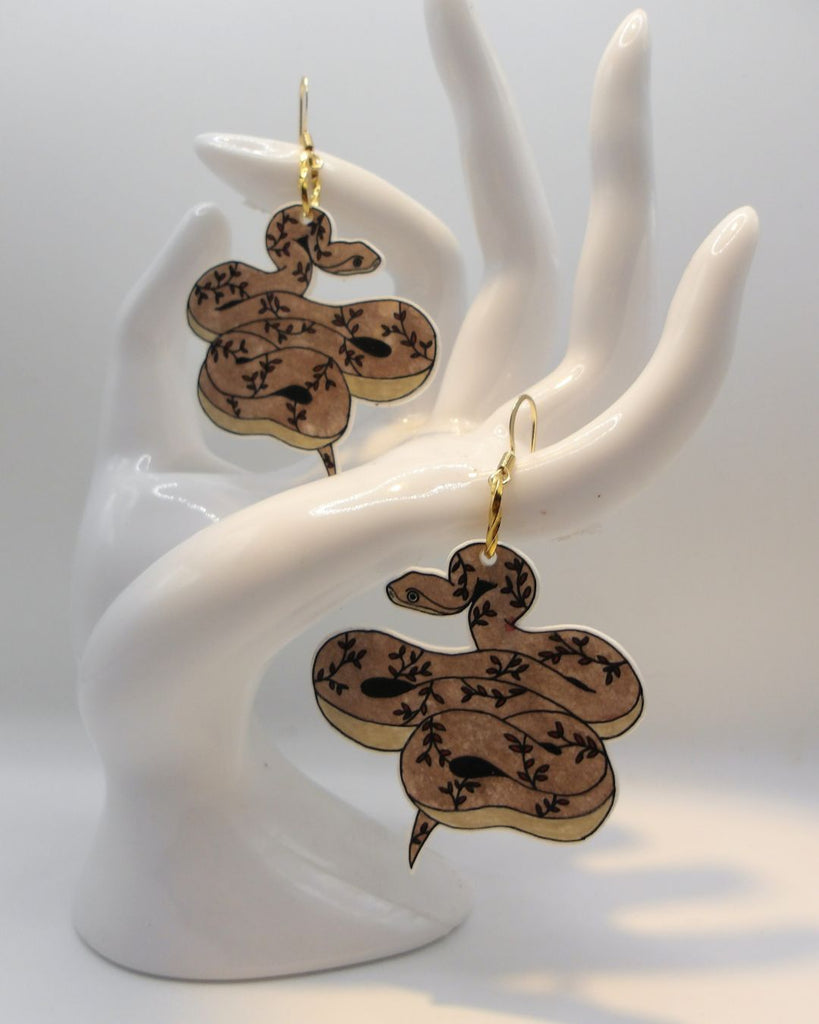 Foto van een paar oorbellen in halloween thema.  De oorbellen hebben goudkleurige hardware, en een tekening van een slangen met takjes in bruin en bordeaux. 