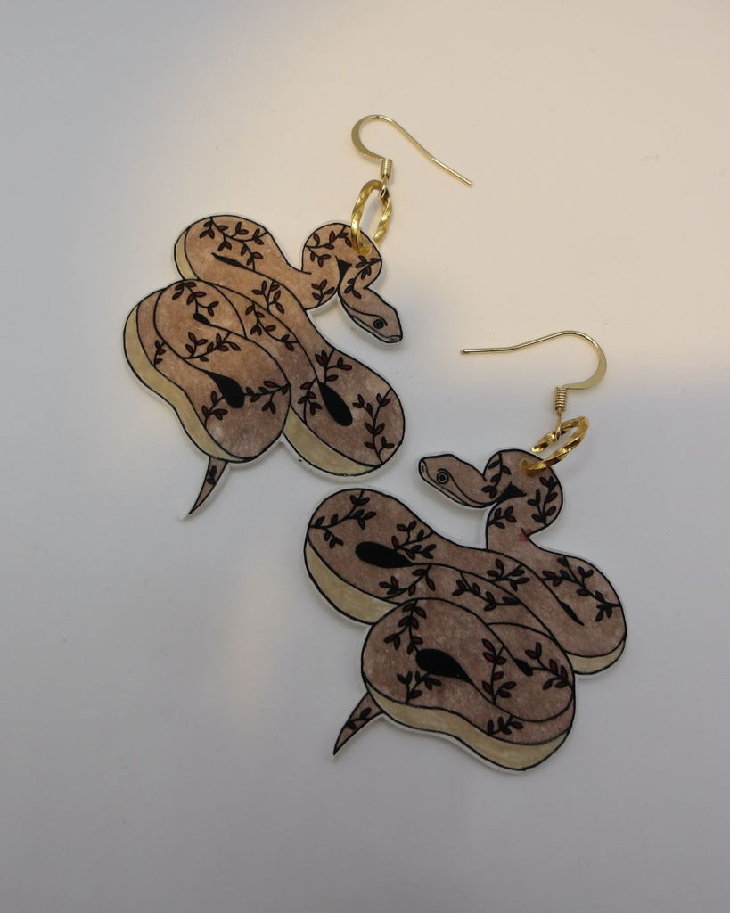 Foto van een paar oorbellen in halloween thema.  De oorbellen hebben goudkleurige hardware, en een tekening van een slangen met takjes in bruin en bordeaux. 