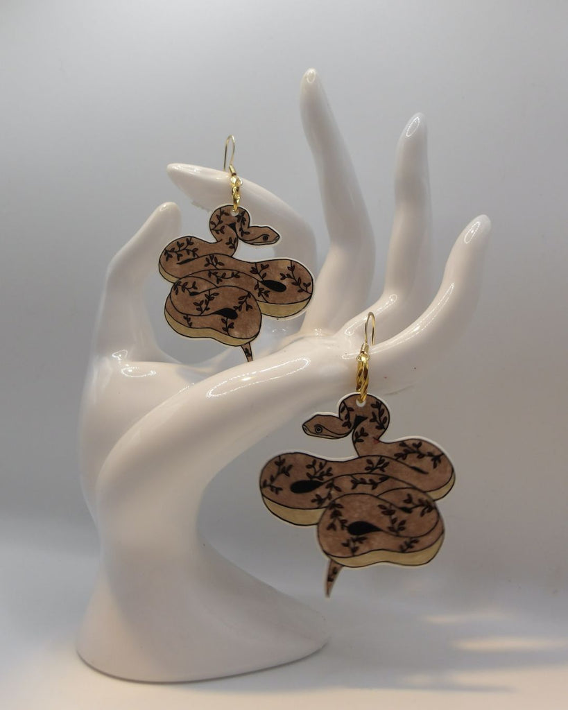 Foto van een paar oorbellen in halloween thema.  De oorbellen hebben goudkleurige hardware, en een tekening van een slangen met takjes in bruin en bordeaux. 