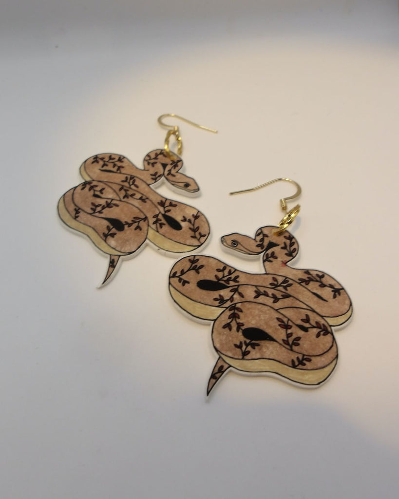 Foto van een paar oorbellen in halloween thema.  De oorbellen hebben goudkleurige hardware, en een tekening van een slangen met takjes in bruin en bordeaux. 