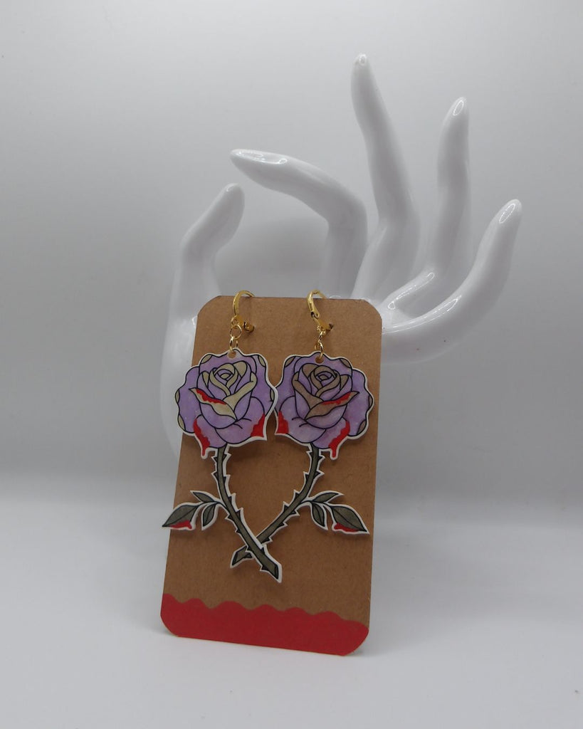 Foto van een paar oorbellen in halloween thema.  De oorbellen hebben goudkleurige hardware, en een tekening van prikende roze rozen. 