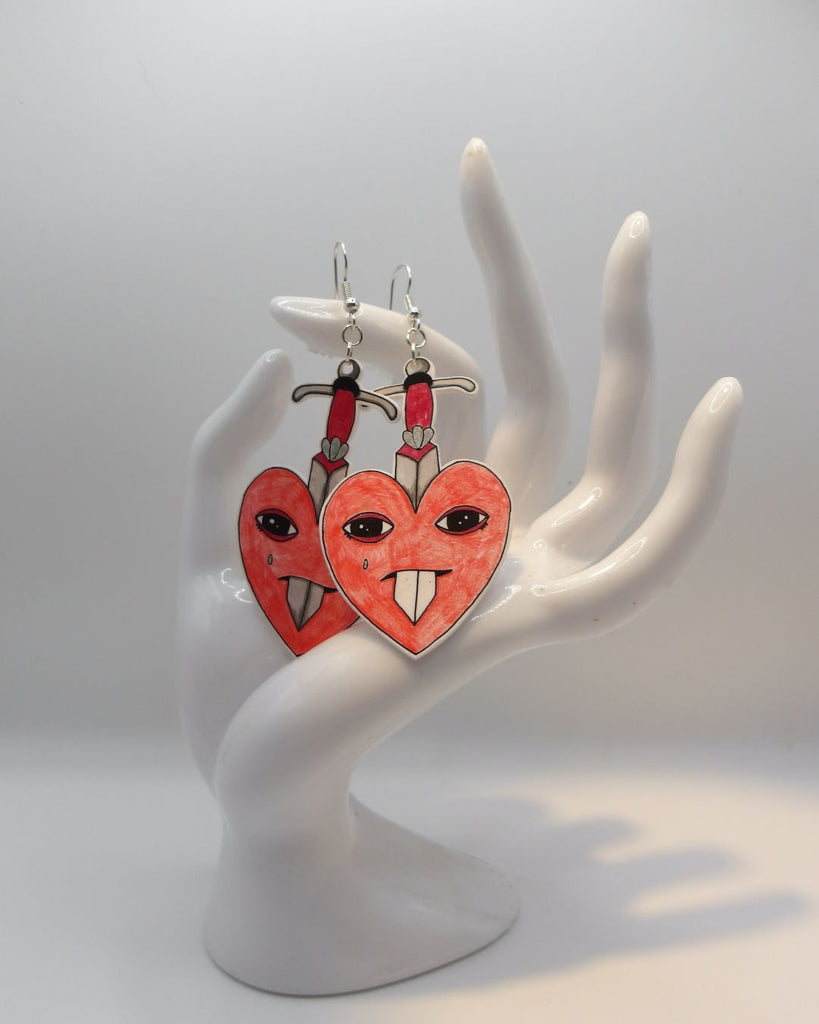 Foto van een paar oorbellen in halloween thema.  De oorbellen hebben zilverkleurige hardware, en een tekening van rode hartjes met een traan. Ze worden doorboord door een zwaard 