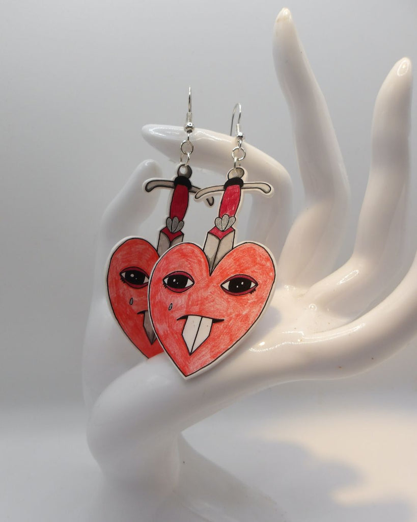 Foto van een paar oorbellen in halloween thema.  De oorbellen hebben zilverkleurige hardware, en een tekening van rode hartjes met een traan. Ze worden doorboord door een zwaard 