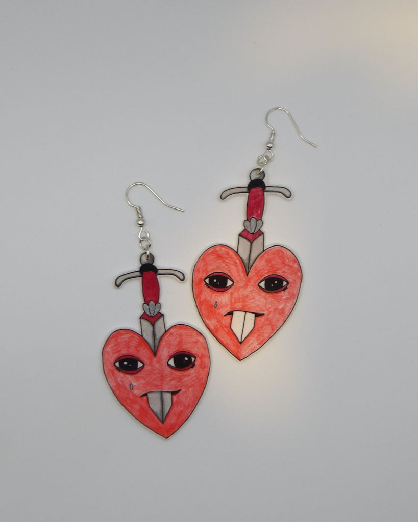 Foto van een paar oorbellen in halloween thema.  De oorbellen hebben zilverkleurige hardware, en een tekening van rode hartjes met een traan. Ze worden doorboord door een zwaard 