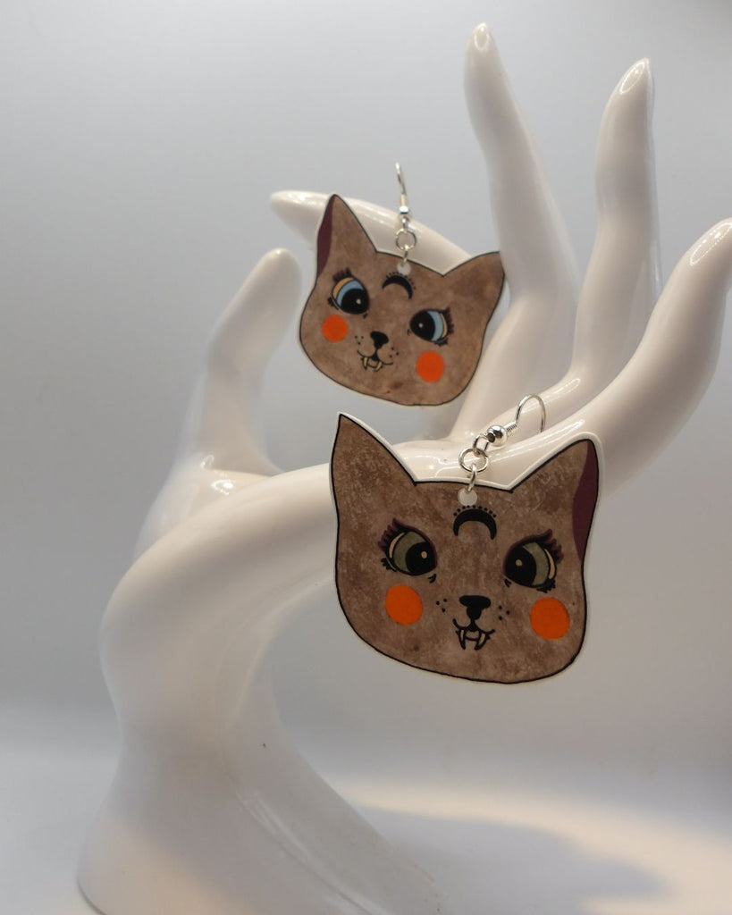 Foto van een paar oorbellen in halloween thema.  De oorbellen hebben zilverkleurige hardware, en een tekening van katten met scherpe hoektanden. De katten zijn bruin met details in oranje, bordeaux en groen