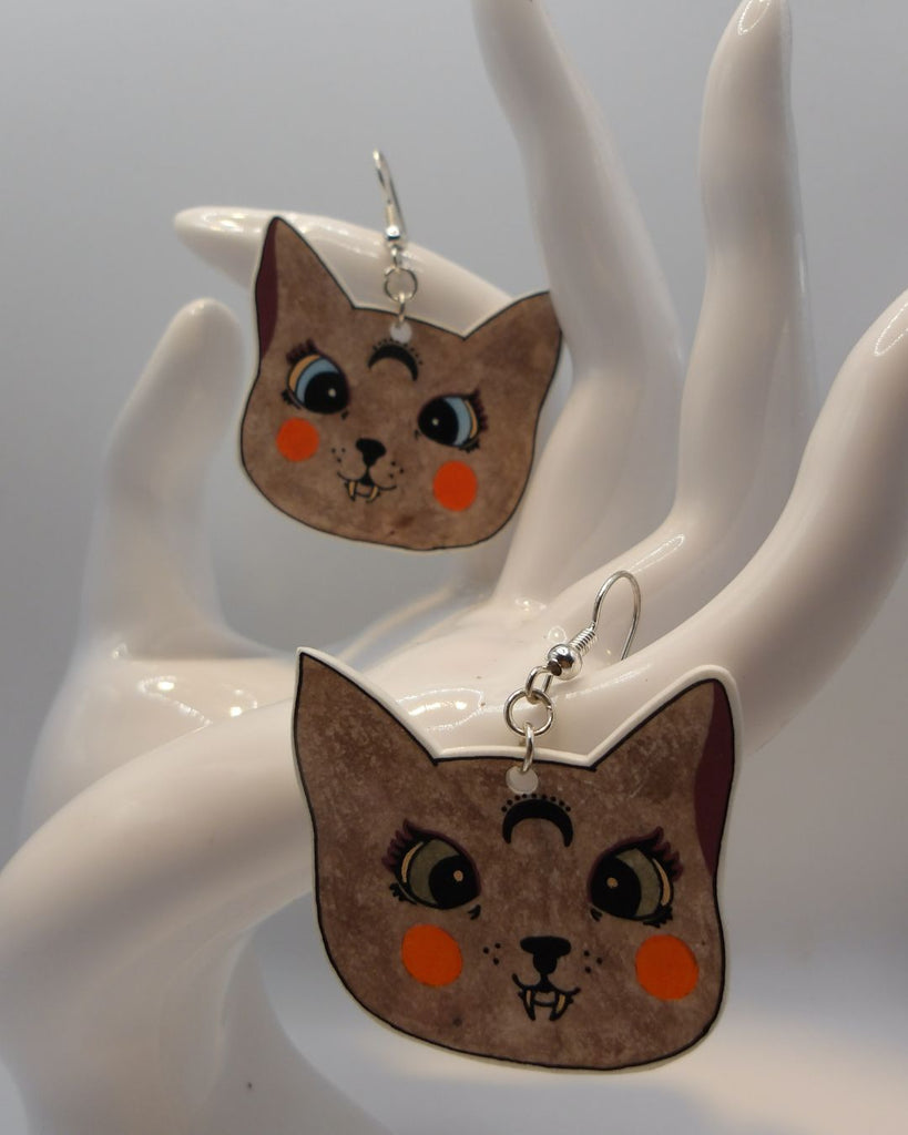 Foto van een paar oorbellen in halloween thema.  De oorbellen hebben zilverkleurige hardware, en een tekening van katten met scherpe hoektanden. De katten zijn bruin met details in oranje, bordeaux en groen