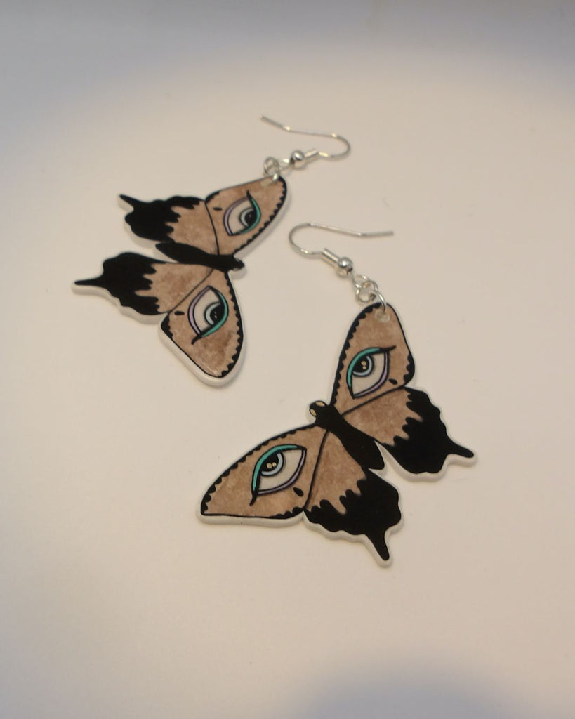 Foto van een paar oobellen in halloween thema.  De oorbellen hebben zilverkleurige hardware, en een tekening van bruine vlinders met huilende ogen en blauwe details. 