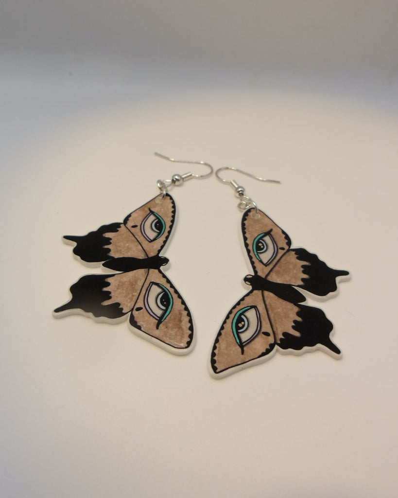 Foto van een paar oobellen in halloween thema.  De oorbellen hebben zilverkleurige hardware, en een tekening van bruine vlinders met huilende ogen en blauwe details. 