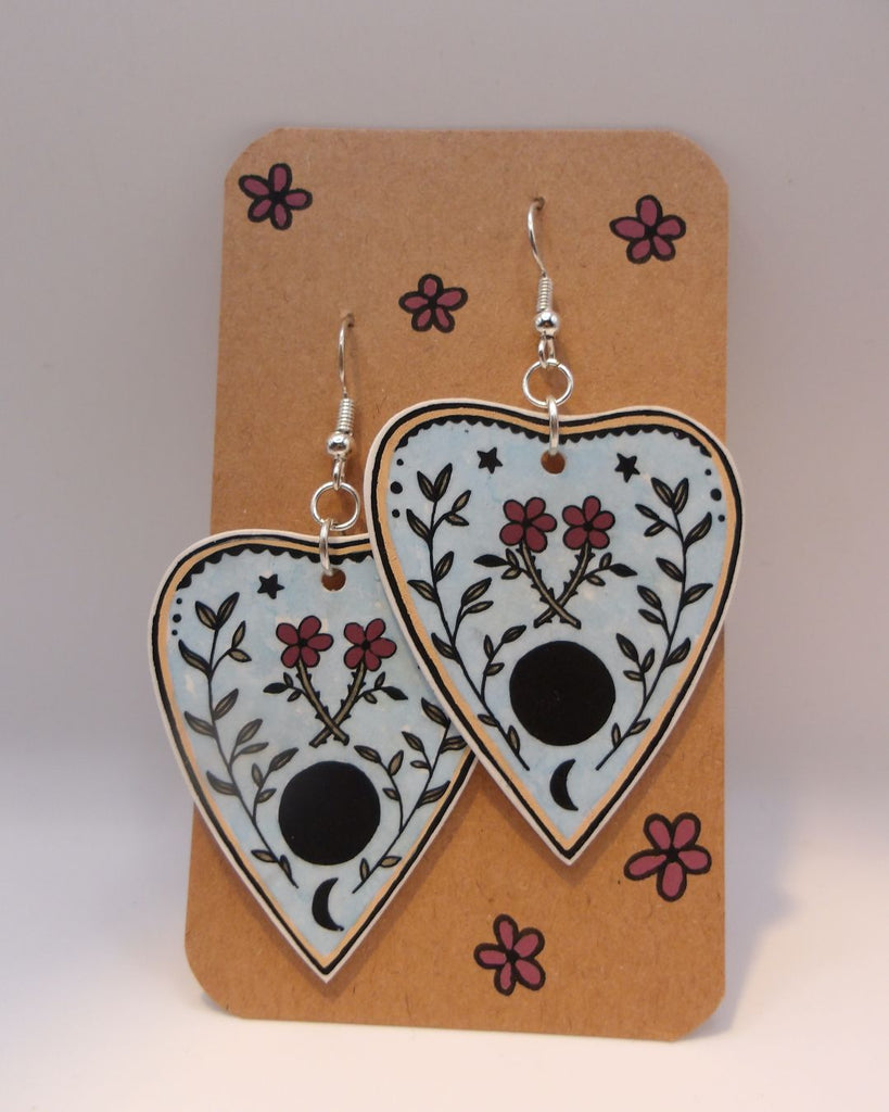 Foto van een paar oorbellen in halloween thema.  De oorbellen hebben zilverkleurige hardware, en een tekening van een ouija spel met bloemen. 