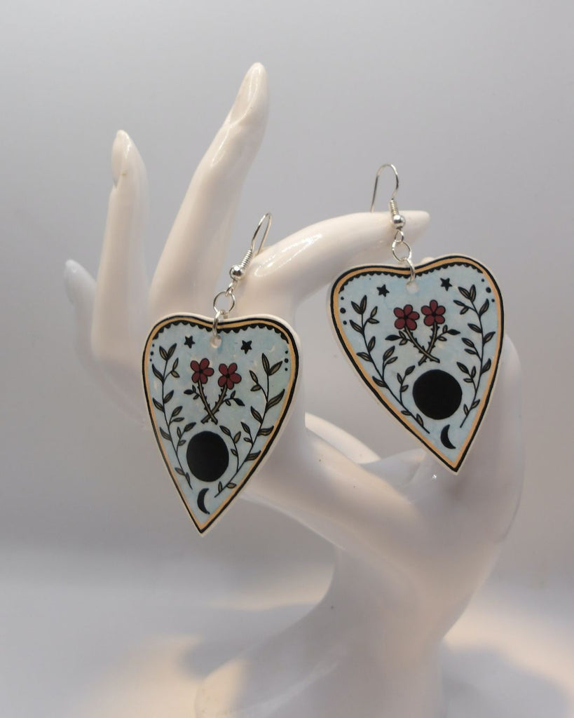 Foto van een paar oorbellen in halloween thema.  De oorbellen hebben zilverkleurige hardware, en een tekening van een ouija spel met bloemen. 