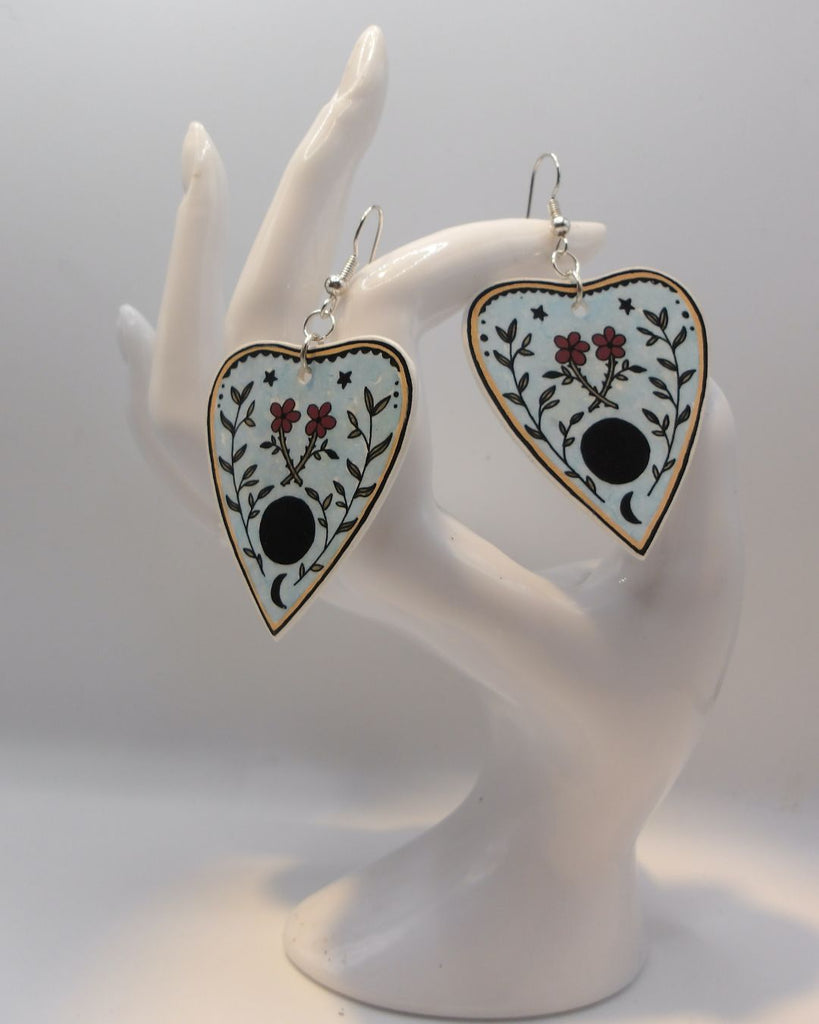 Foto van een paar oorbellen in halloween thema.  De oorbellen hebben zilverkleurige hardware, en een tekening van een ouija spel met bloemen. 