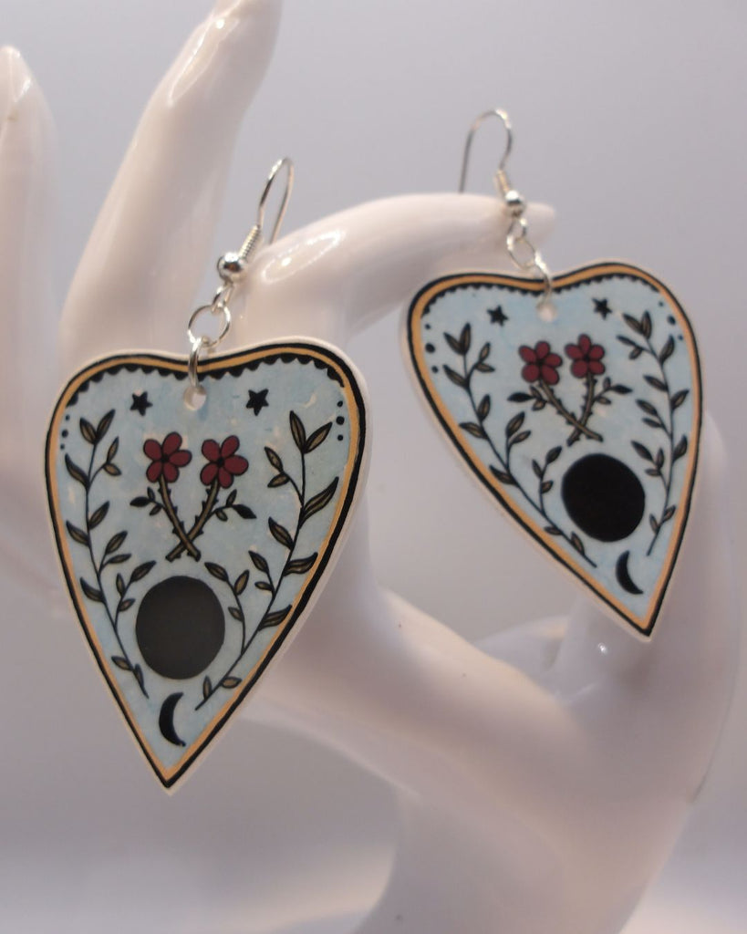 Foto van een paar oorbellen in halloween thema.  De oorbellen hebben zilverkleurige hardware, en een tekening van een ouija spel met bloemen. 