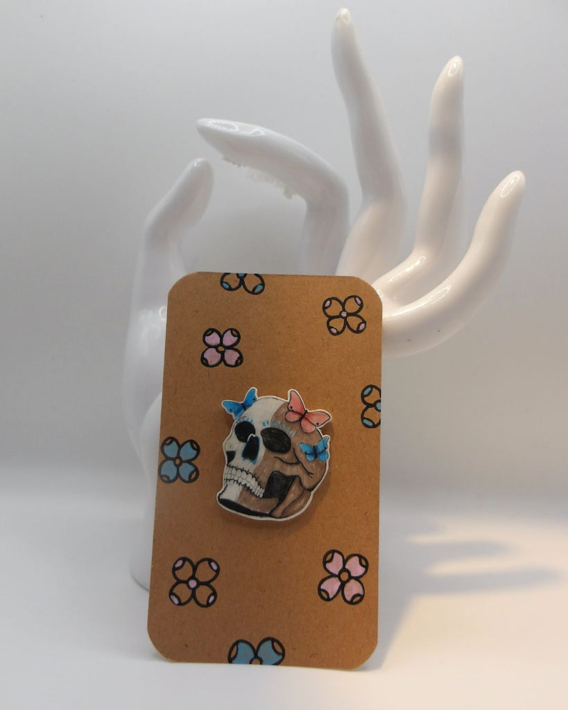 Foto van een pin in halloween thema.  De pin toont een doodshoofd met 3 kleurrijke vlinders op zijn hoofd. in kleuren beige, blauw en roze. 