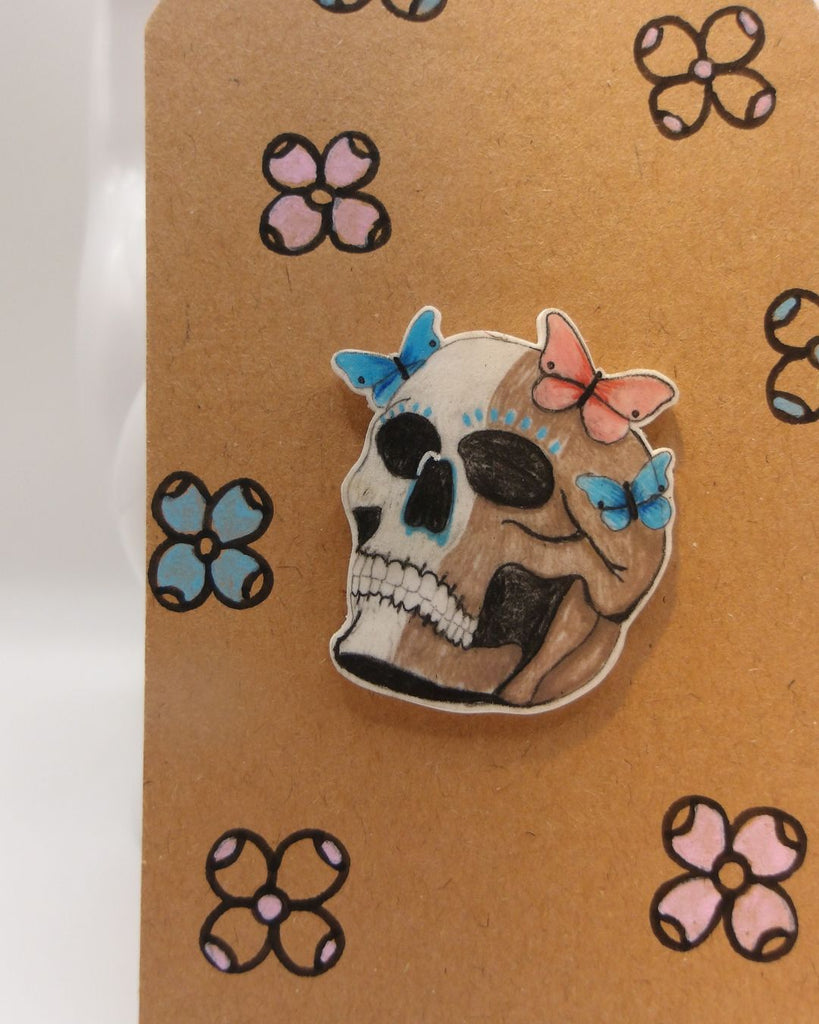 Foto van een pin in halloween thema.  De pin toont een doodshoofd met 3 kleurrijke vlinders op zijn hoofd. in kleuren beige, blauw en roze. 