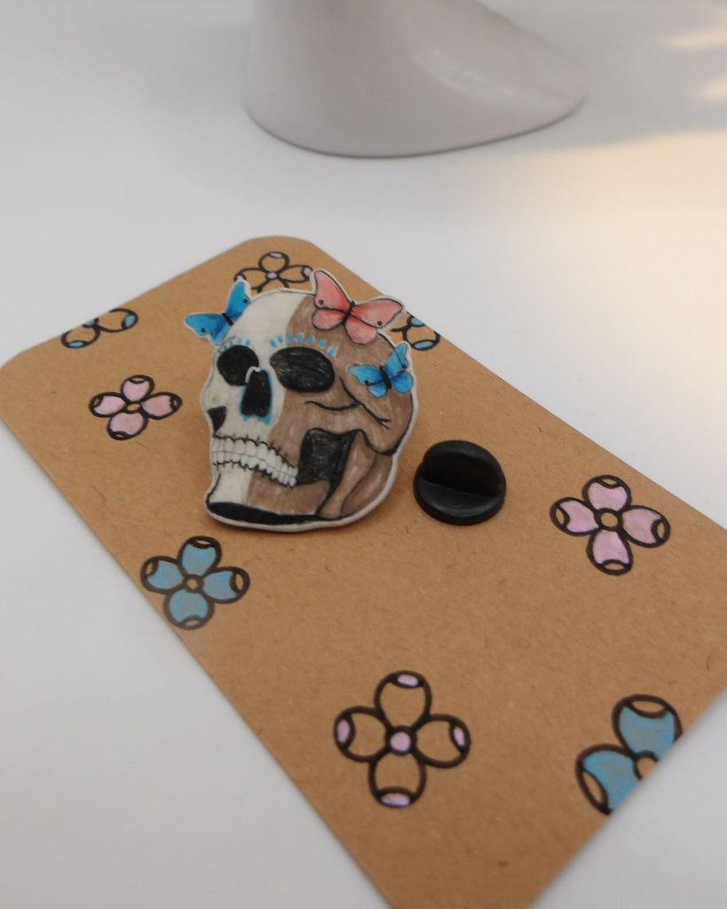 Foto van een pin in halloween thema.  De pin toont een doodshoofd met 3 kleurrijke vlinders op zijn hoofd. in kleuren beige, blauw en roze. 
