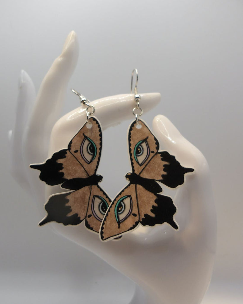 Foto van een paar oobellen in halloween thema.  De oorbellen hebben zilverkleurige hardware, en een tekening van bruine vlinders met huilende ogen en blauwe details. 