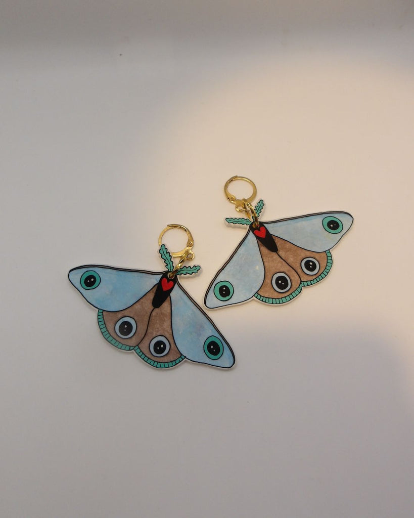 Foto van een paar oorbellen in halloween thema.  De oorbellen hebben goudkleurige hardware, en een tekening van een blauwe mot met een rood hartje. 