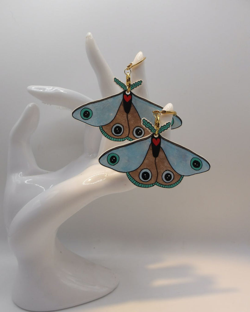 Foto van een paar oorbellen in halloween thema.  De oorbellen hebben goudkleurige hardware, en een tekening van een blauwe mot met een rood hartje. 