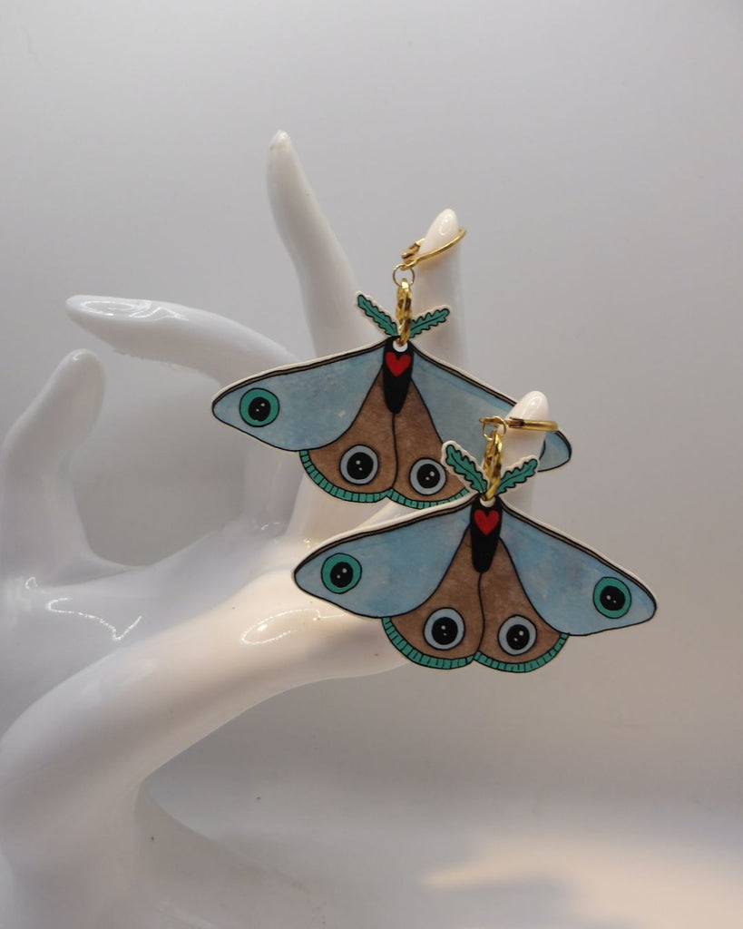 Foto van een paar oorbellen in halloween thema.  De oorbellen hebben goudkleurige hardware, en een tekening van een blauwe mot met een rood hartje. 