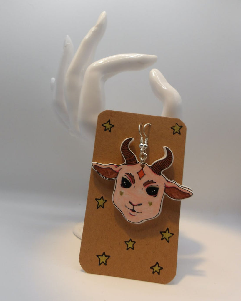 Foto van een paar oobellen in halloween thema. De oorbellen zijn gepresenteerd op een kaartje met sterretjes. De oorbellen hebben zilverkleurige hardware, en een tekening van een schattig duivels geitje met grote oren en hoorns in het roze.