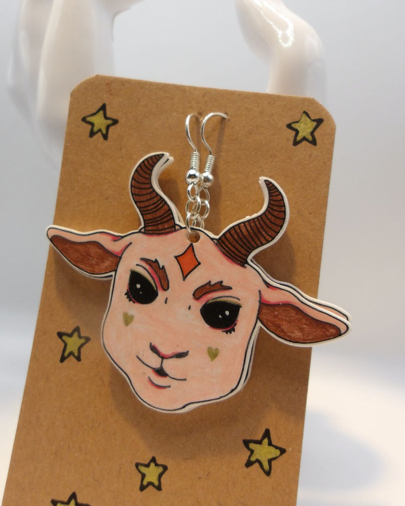 Ingezoomde foto van een paar oobellen in halloween thema. De oorbellen zijn gepresenteerd op een kaartje met sterretjes. De oorbellen hebben zilverkleurige hardware, en een tekening van een schattig duivels geitje met grote oren en hoorns in het roze.