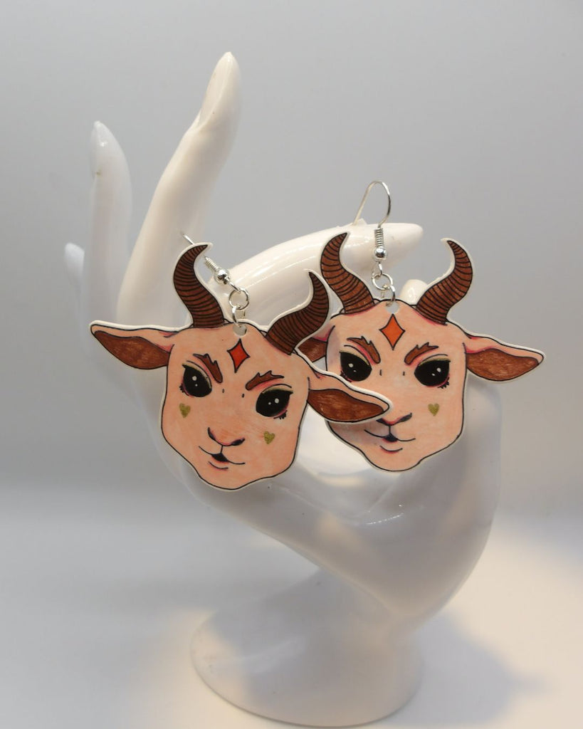 Foto van een paar oobellen in halloween thema. De oorbellen zijn gepresenteerd op een kaartje met sterretjes. De oorbellen hebben zilverkleurige hardware, en een tekening van een schattig duivels geitje met grote oren en hoorns in het roze.