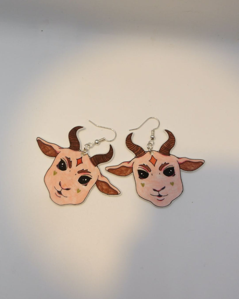 Foto van een paar oobellen in halloween thema. De oorbellen zijn gepresenteerd op een kaartje met sterretjes. De oorbellen hebben zilverkleurige hardware, en een tekening van een schattig duivels geitje met grote oren en hoorns in het roze.