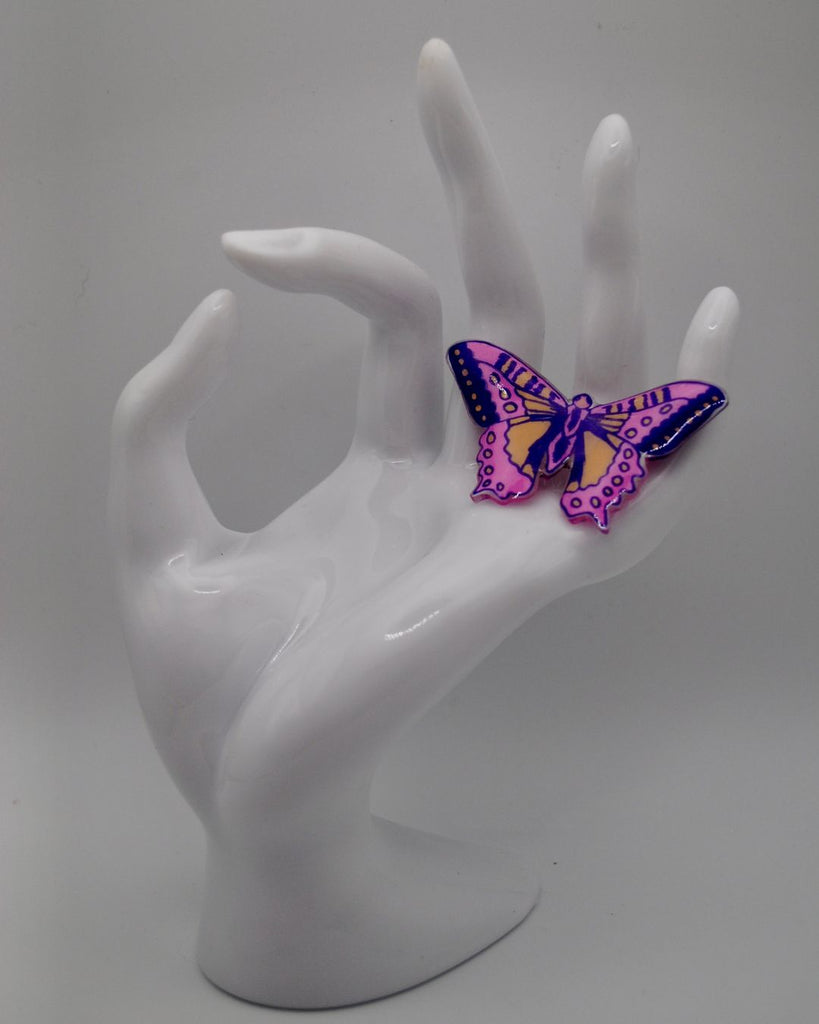 Foto van een unieke statement ring met vlinder. De ring is met de hand getekend. De vlinder is roze met paars en peach. Op deze foto wordt de ring gepresenteerd aan een witte hand en tegen een witte achtergrond. 