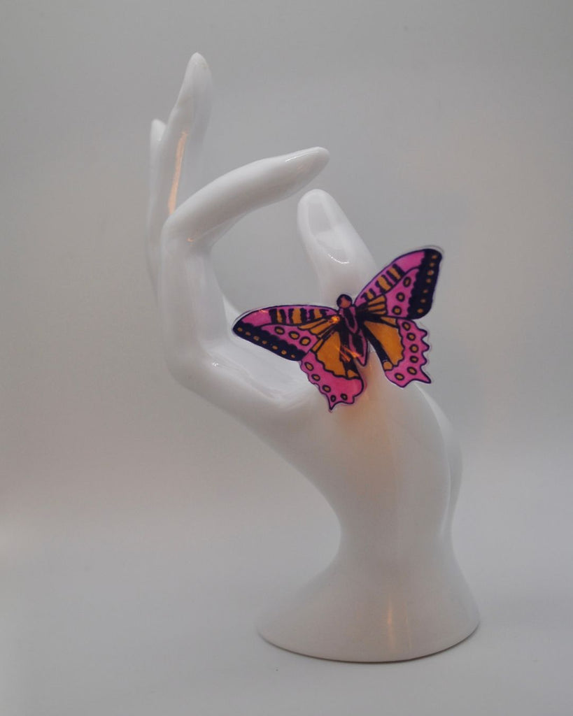 Foto van een unieke statement ring met vlinder. De ring is met de hand getekend. De vlinder is roze met paars en peach. Op deze foto wordt de ring gepresenteerd aan een witte hand en tegen een witte achtergrond.  Ook is er een mooi licht aanwezig