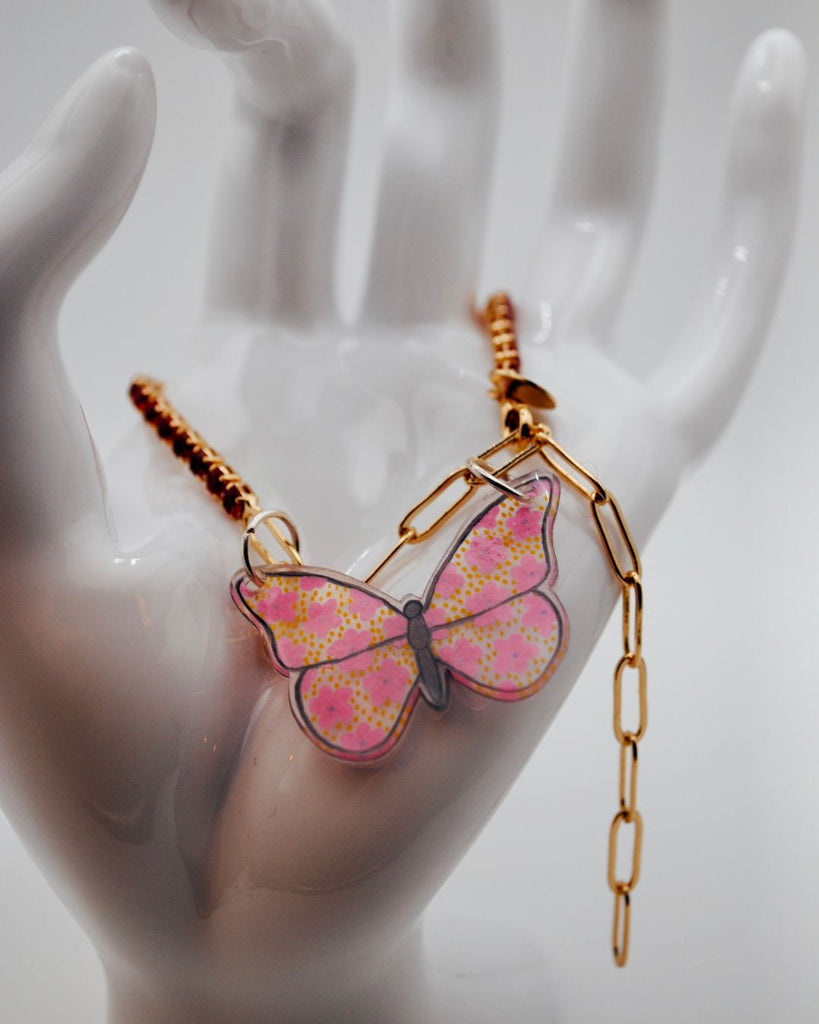 Detailfoto van armbandje met rode steentjes en roze vlinder in krimpfolie. De vlinder heeft roze bloemetjes met gele stippen