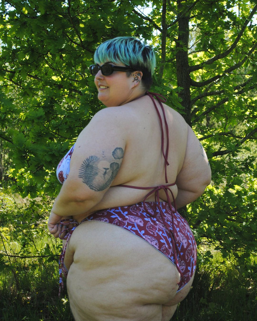 Achteraanzicht. Het model draagt een mismatch bikini top en broekje. De bikini heeft een bloemenprint. Het topje is helder blauw en het broekje is bordeaux.