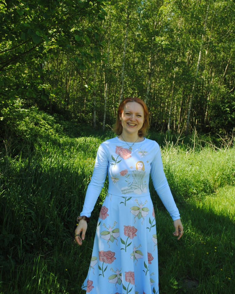 Het lachende model draagt een zomers blauw jurkje met zakken en lange mouwen. Het zwierige jurkje komt tot onder de knie. Het heeft een bloemenprint met een meisje dat tussen de bloemetjes zit. 