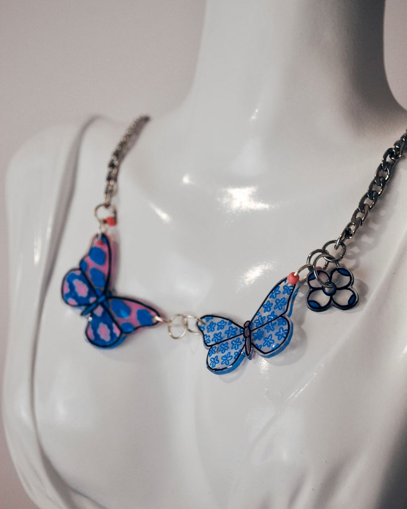 Foto van zilverkleurige ketting met 2 vlinder hangers . De linkste vlinder is roze met blauwe wolkjes. De rechtse vlinder is doorzichtig met een blauwe bloemetjes motief 