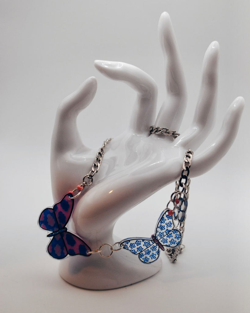 Foto van zilverkleurige ketting  gedrapeerd over hand. De ketting heeft 2 vlinder hangers . De linkste vlinder is roze met blauwe wolkjes. De rechtse vlinder is doorzichtig met een blauwe bloemetjes motief 