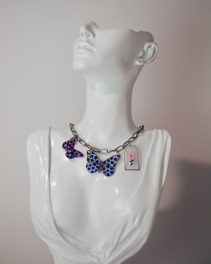 Foto van zilverkleurige ketting met 2 vlinder hangers en 1 tag met een bloem. De vlinders uit krimpfolie zijn blauw en blauw met roze 