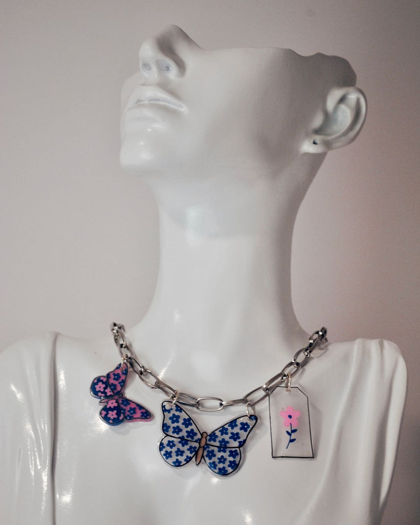 Foto van zilverkleurige ketting met 2 vlinder hangers en 1 tag met een bloem. De vlinders uit krimpfolie zijn blauw en blauw met roze 