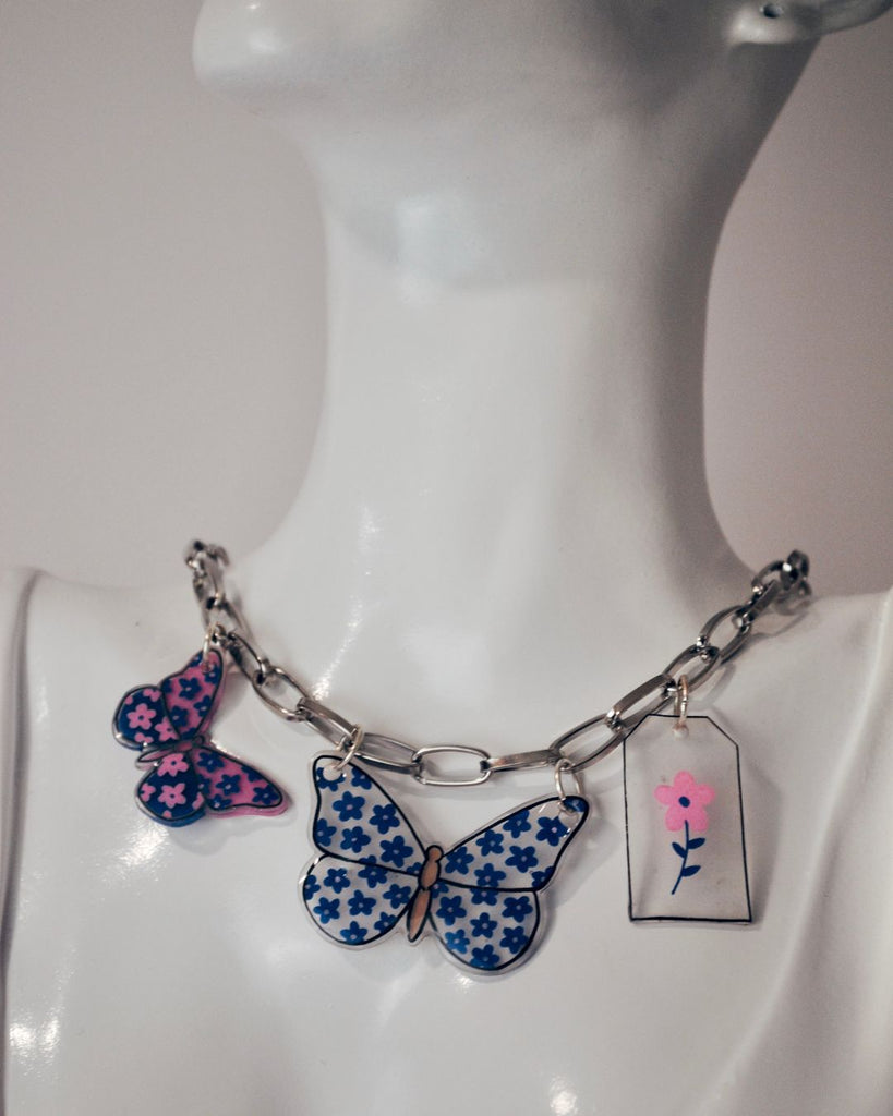 Foto dichtbij van zilverkleurige ketting met 2 vlinder hangers en 1 tag met een bloem. De vlinders uit krimpfolie zijn blauw en blauw met roze 
