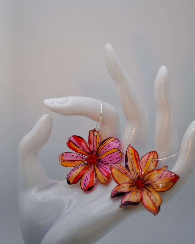 Detail foto van unieke bloemen oorbellen. Ze worden gepresenteerd tegen een witte achtergrond. Ze hangen aan een witte hand. De oorbellen zijn 2 verschillende bloemen in roze, geel en oranje. De bloemen hangen aan kleine ,dunne oorringetjes 