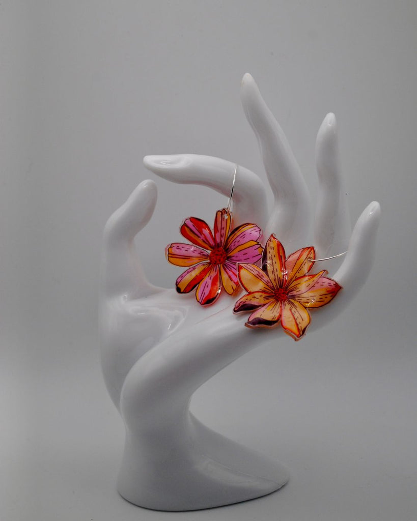  Foto van verder van unieke bloemen oorbellen. Ze worden gepresenteerd tegen een witte achtergrond. Ze hangen aan een witte hand. De oorbellen zijn 2 verschillende bloemen in roze, geel en oranje. De bloemen hangen aan kleine ,dunne oorringetjes 