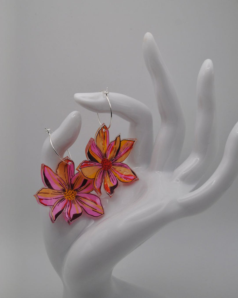 Foto van een paar bloemen oorbellen. De oorbellen zijn 2 met de hand getekende (bijna identieke) bloemen uit krimpfolie. Ze zijn roze met geel en oranje details. Ze worden gepresenteerd hangend aan een wit beeld van een hand, tegen een witte achtergrond 