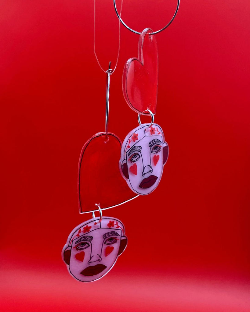 Foto tegen rode achtergrond. Op de voorgrond hangen 2 oorbellen.  Het  zijn lange hangers bestaande uit een groot hart met daaronder een roze gezichtje met hartjes wangen